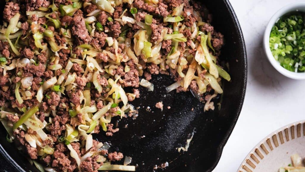 Ground beef and cabbage stir-fried in a skillet, with chopped green onions in a bowl beside it.