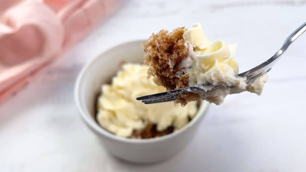 A fork holding a piece of cake with cream cheese frosting over a frosted cake mix mug cake on a table.