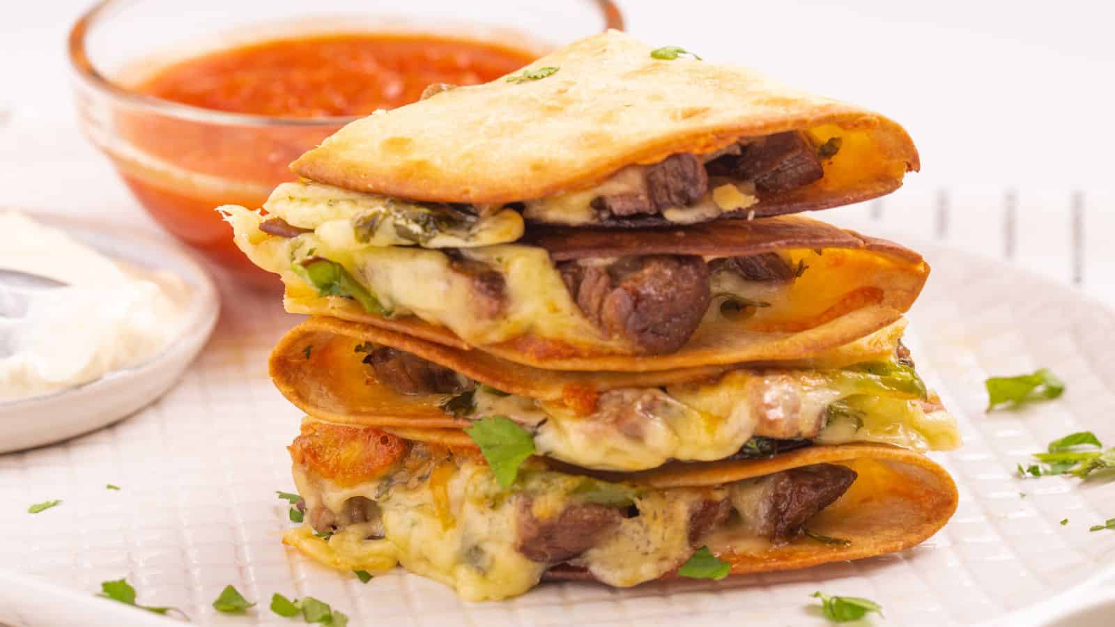 A stack of quesadillas filled with cheese and steak served with a side of red salsa on a white plate.