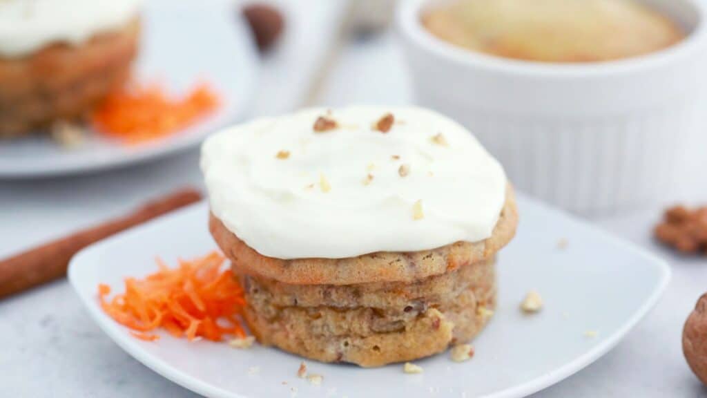 A carrot cake muffin topped with cream cheese frosting and chopped nuts, served on a white plate with grated carrot on the side.