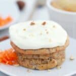 A carrot cake muffin topped with cream cheese frosting and chopped nuts, served on a white plate with grated carrot on the side.