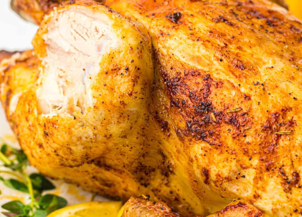 Close-up of a roasted chicken with crispy, golden-brown skin, seasoned with herbs, served with lemon slices.