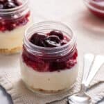 Two jars of layered dessert with a cheesecake base, a middle layer of red fruit compote, and topped with whole cherries, served on a cloth with spoons nearby.