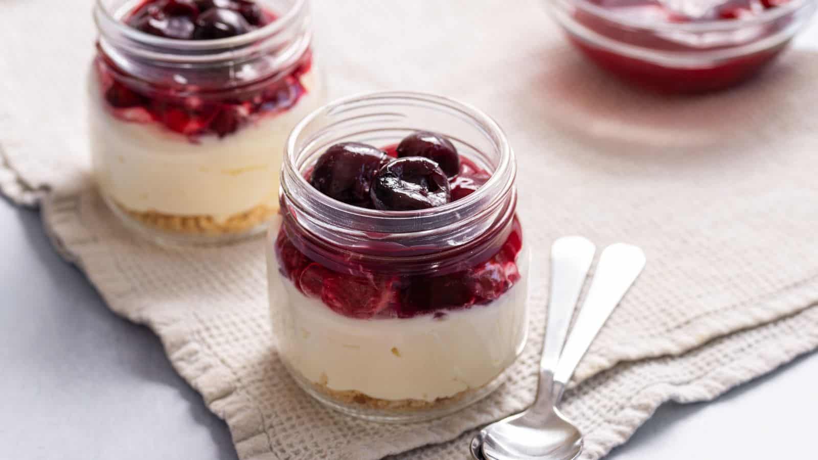 Two jars of layered cheesecake with a graham cracker base, cream cheese filling, and a topping of dark red cherries, on a cloth with spoons beside them.