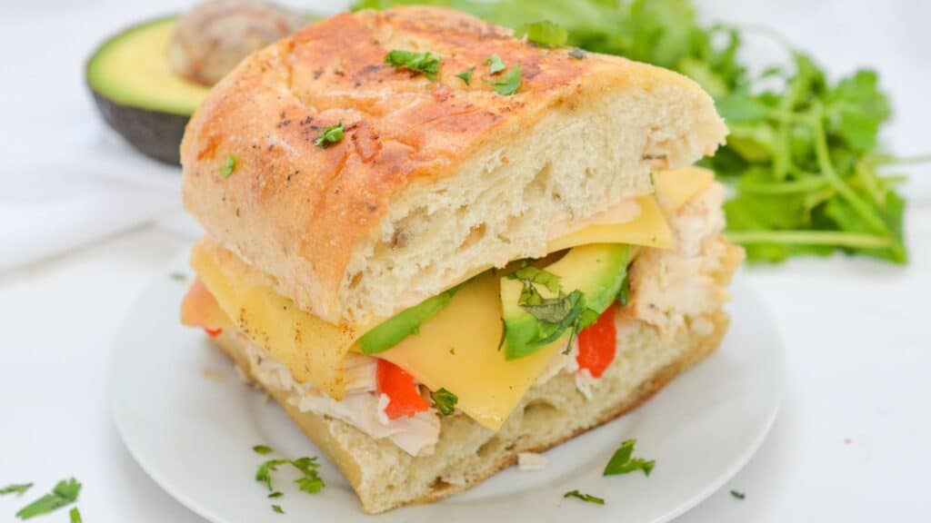 A fresh sandwich with chicken, cheese, avocado on a white plate.