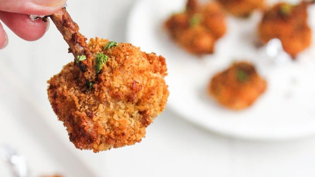 A close-up image of a hand holding a crispy, breaded chicken drumstick garnished with herbs, with more drumsticks on a plate in the background.