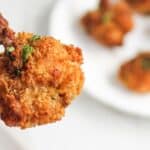 A close-up image of a hand holding a crispy, breaded chicken drumstick garnished with herbs, with more drumsticks on a plate in the background.