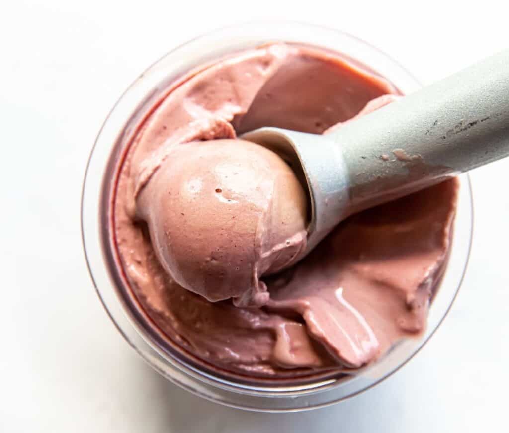A scoop of creamy strawberry ice cream being lifted out of a glass container with a metal spoon.