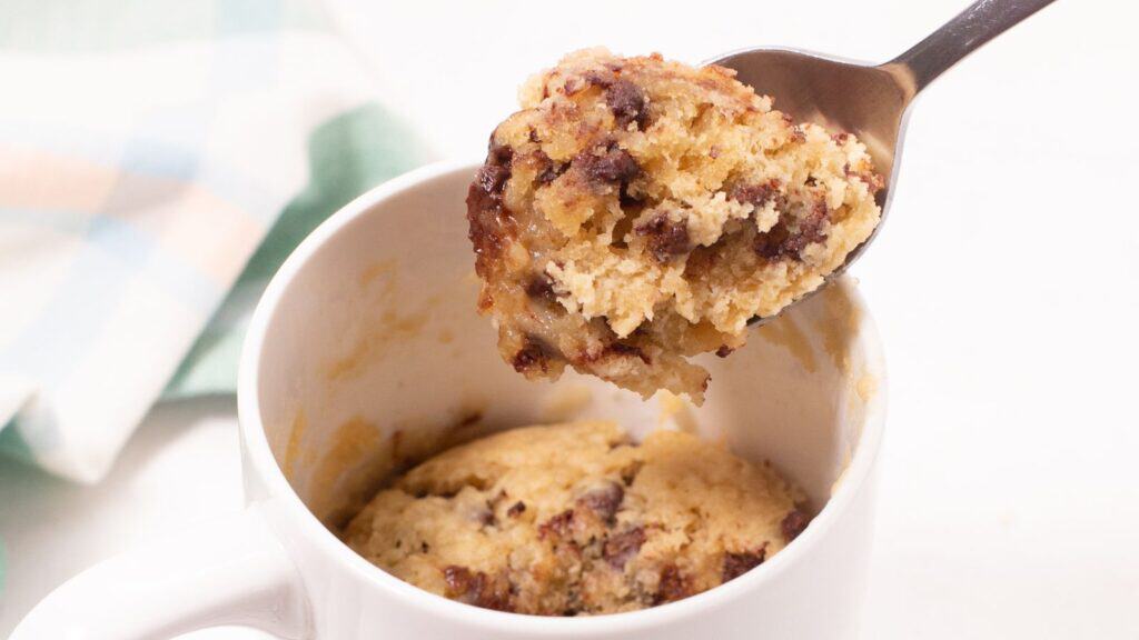 A spoon lifting a portion of a freshly microwaved chocolate chip mug cake from a white ceramic mug.