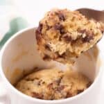 A spoon lifting a portion of a freshly microwaved chocolate chip mug cake from a white ceramic mug.