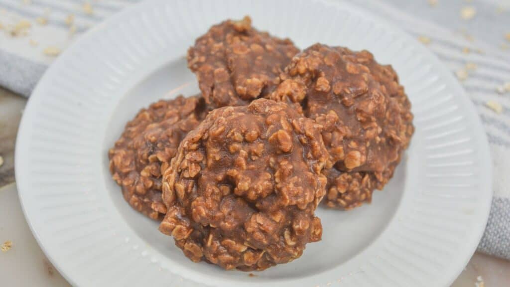 Three no-bake chocolate oatmeal cookies on a white plate with crumbs scattered around, on a light-colored surface.