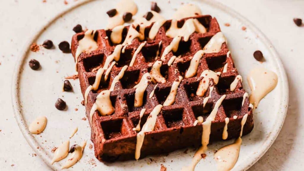 A chocolate waffle drizzled with cream sauce on a plate, garnished with chocolate chips and powdered chocolate.