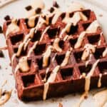 A chocolate waffle drizzled with cream sauce on a plate, garnished with chocolate chips and powdered chocolate.