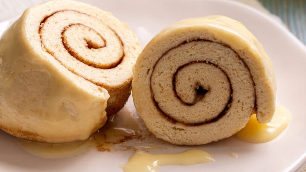 Two slices of cinnamon roll cake with creamy icing on a white plate, drizzled with extra icing.