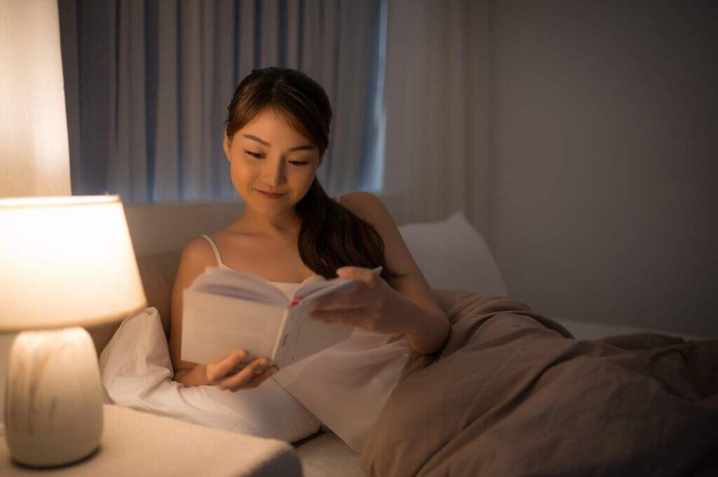 Woman reading a book in bed by lamp light, aligning with her circadian rhythm.