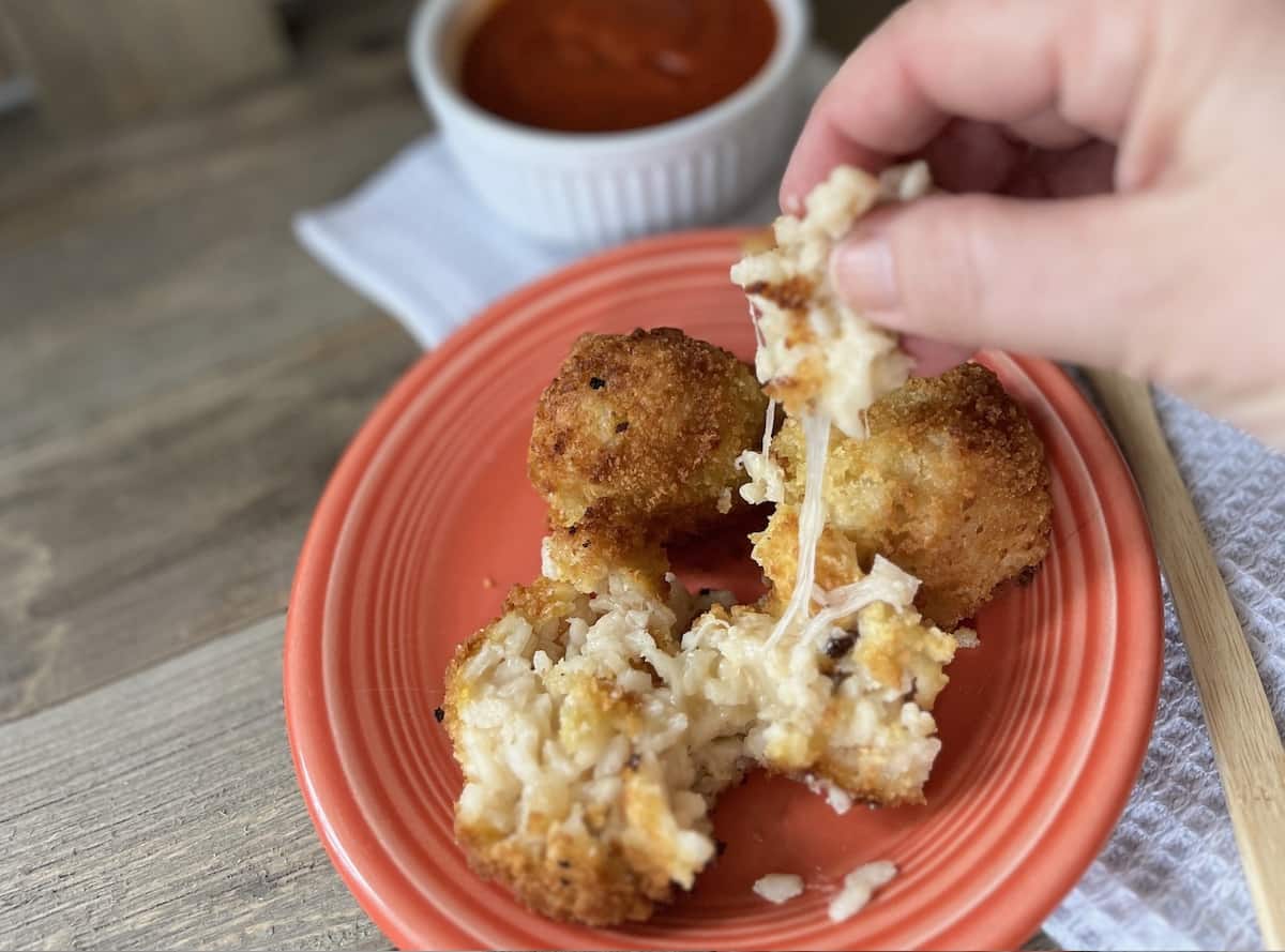 A person pulling apart a gooey, cheese-filled arancini with more of the fried rice balls and sauce in the background.
