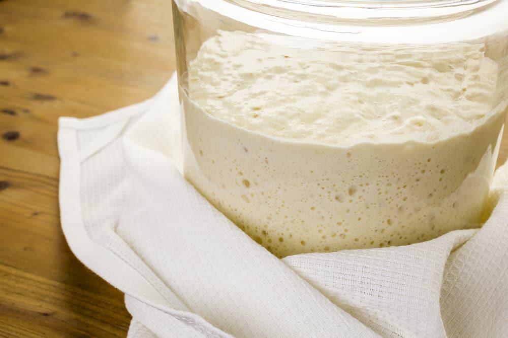 A glass jar filled with active sourdough starter ready for feeding sits on a wooden surface.