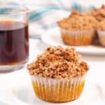 A cup of black coffee with two streusel-topped muffins on a white plate, with a striped napkin in the background.