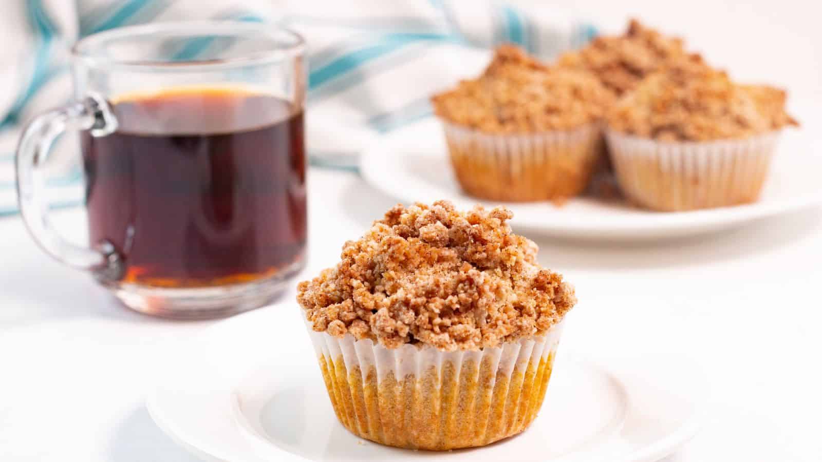 A coffee cake muffin with a crumbly topping on a white plate, next to a glass of black coffee and another muffin in the background.