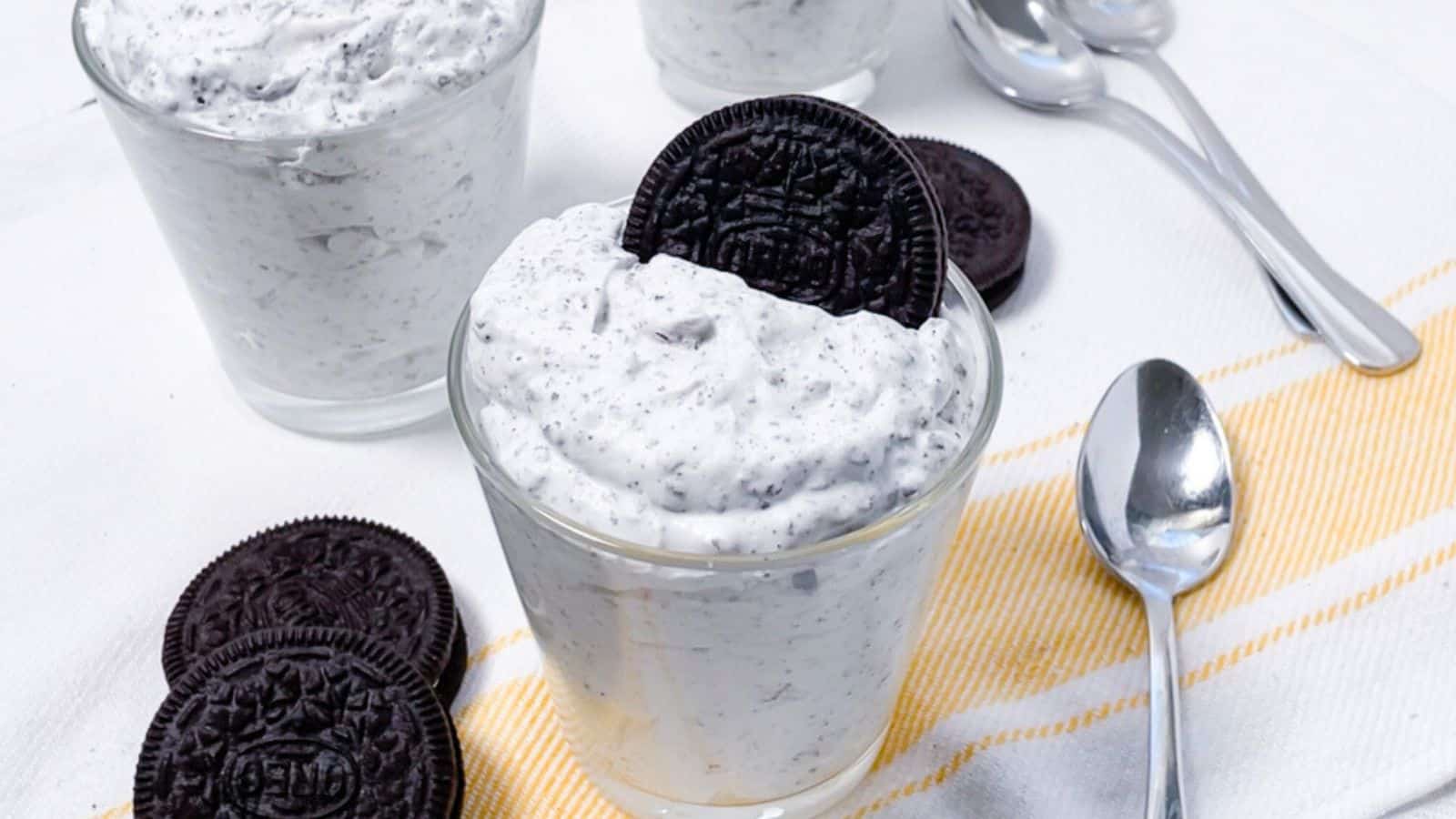 Two glasses of cookies and cream dessert topped with oreo cookies, served on a white tablecloth with a yellow-striped napkin and spoons.