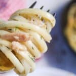 A close-up of spaghetti twirled on a fork with bits of crab meat and herbs, with more pasta and a dish in the blurred background.