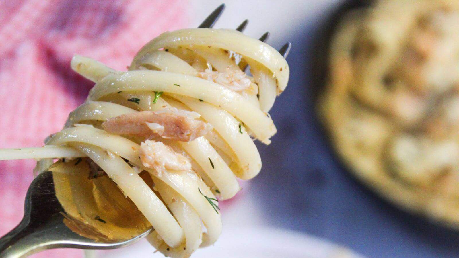 A close-up of spaghetti twirled on a fork with bits of crab meat and herbs, with more pasta and a dish in the blurred background.