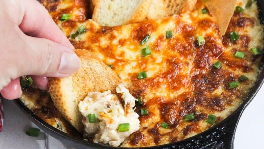 A person dipping a slice of bread into a cheesy dip in a cast-iron skillet, garnished with green onions.