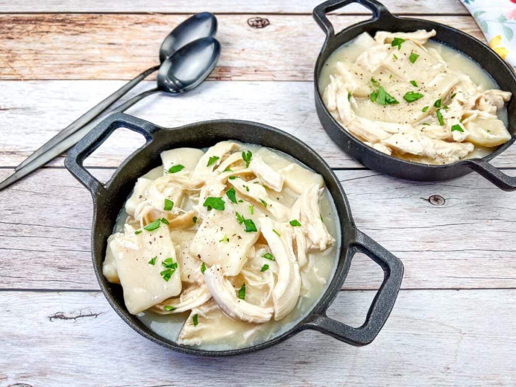 Two bowls of chicken and dumplings garnished with chopped herbs on a wooden table, with spoons on the side.