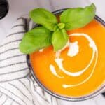 A bowl of creamy tomato soup garnished with fresh basil, viewed from above next to a striped napkin and a pair of sunglasses.