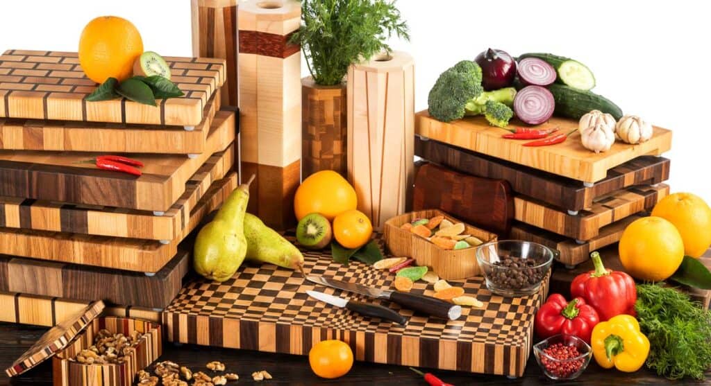 Various wooden cutting boards and kitchen accessories surrounded by fresh fruits, vegetables, and nuts on a white background.