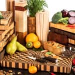 Various wooden cutting boards and kitchen accessories surrounded by fresh fruits, vegetables, and nuts on a white background.