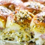 Baked sliders with melted cheese and ham on poppy seed-topped buns in a baking dish.
