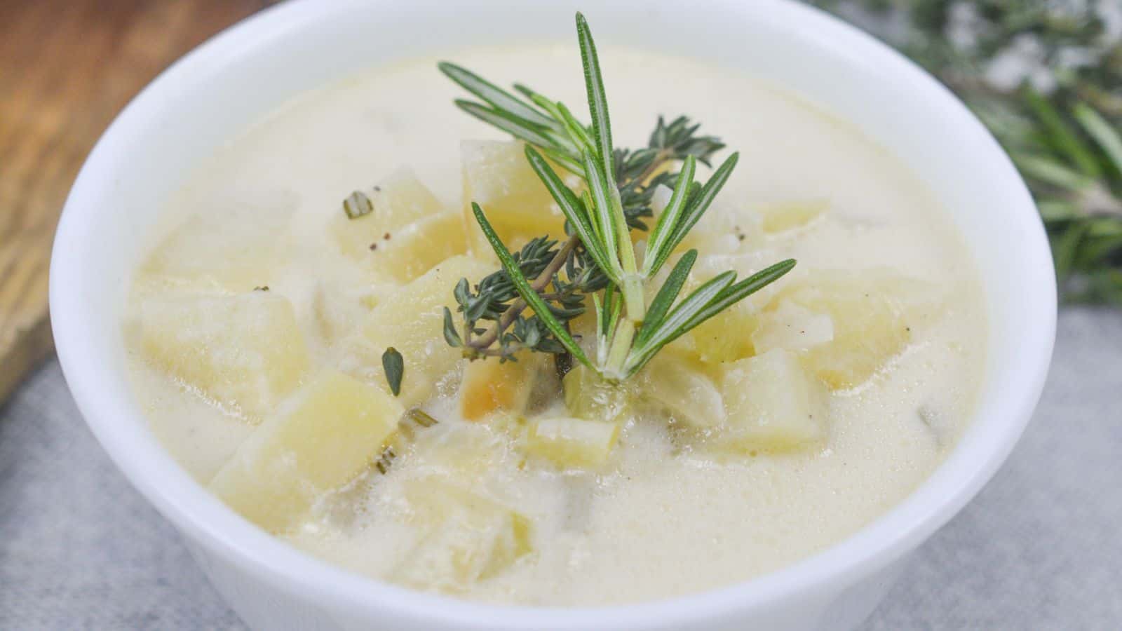 A bowl of creamy potato soup garnished with rosemary and thyme, served on a wooden table.