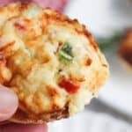 A close-up of a hand holding an egg white frittata muffin with visible toppings of cheese and chopped peppers.