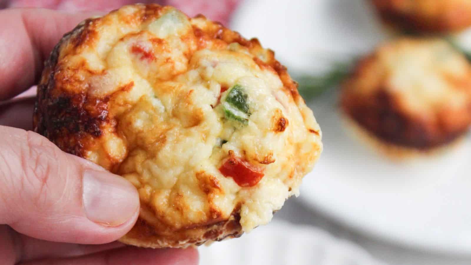 A close-up of a hand holding an egg white frittata muffin with visible toppings of cheese and chopped peppers.