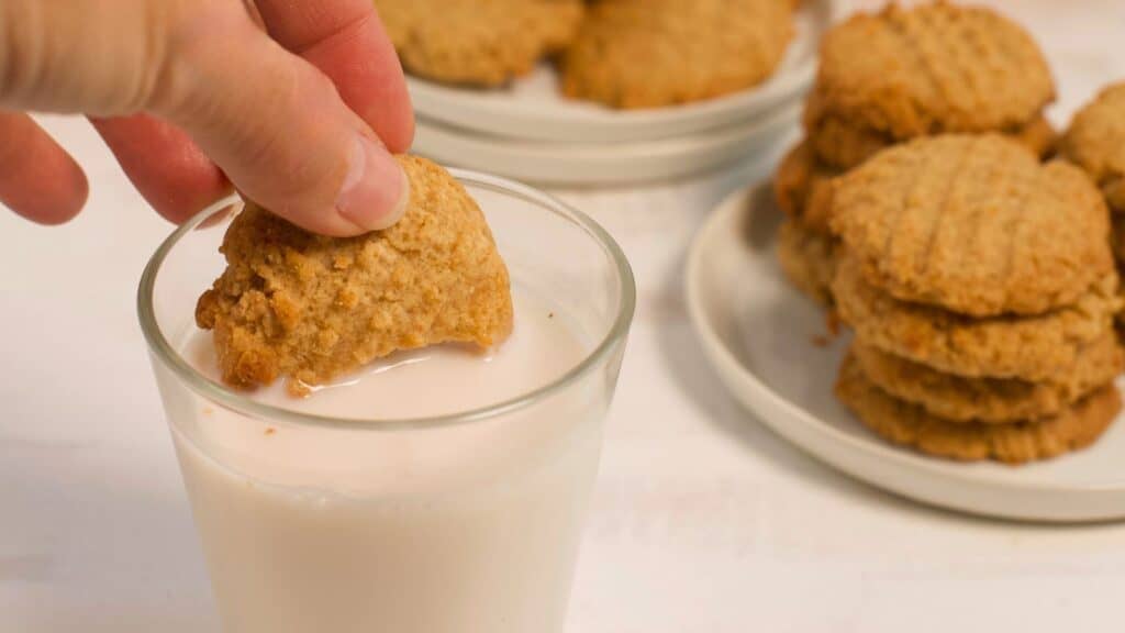 A person dipping a peanut butter cookie into a glass of milk, with more stacked cookies on a plate in the background.