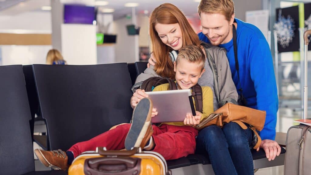 A smiling family sits together in an airport terminal, watching something on a tablet. The child is seated between the two adults, with a suitcase in the foreground. They seem relaxed and happy, perhaps researching their next vacation destination and wondering, 