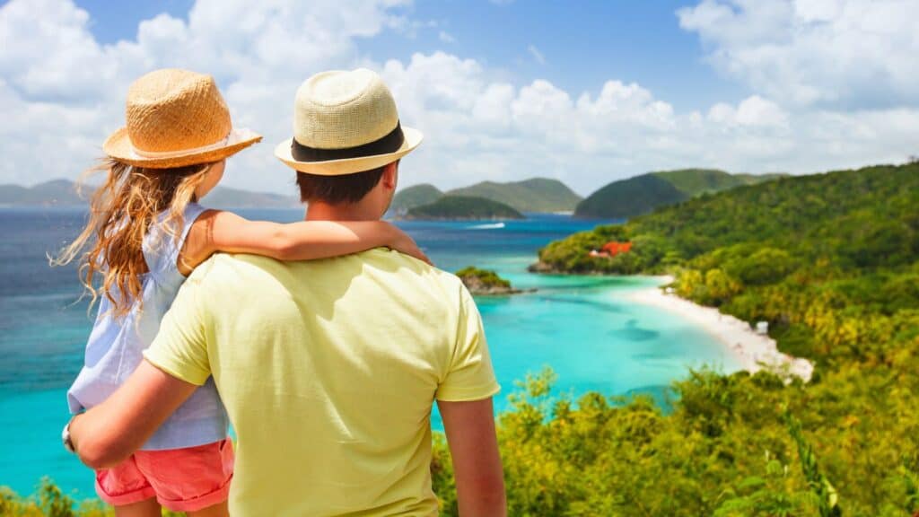 A couple wearing hats and casual summer clothes enjoying a scenic view of a tropical beach and lush greenery.