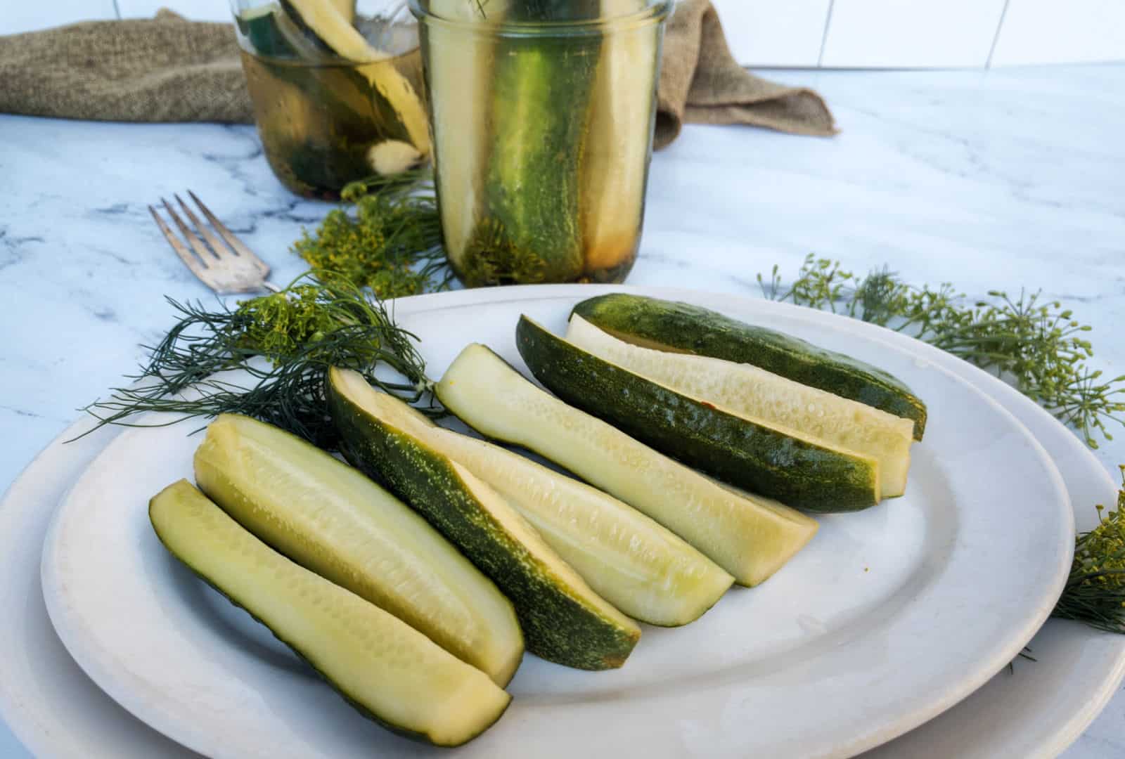 Sliced pickles on a white plate, with whole pickles in a jar and fresh dill on a table, suggesting a homemade pickling theme.