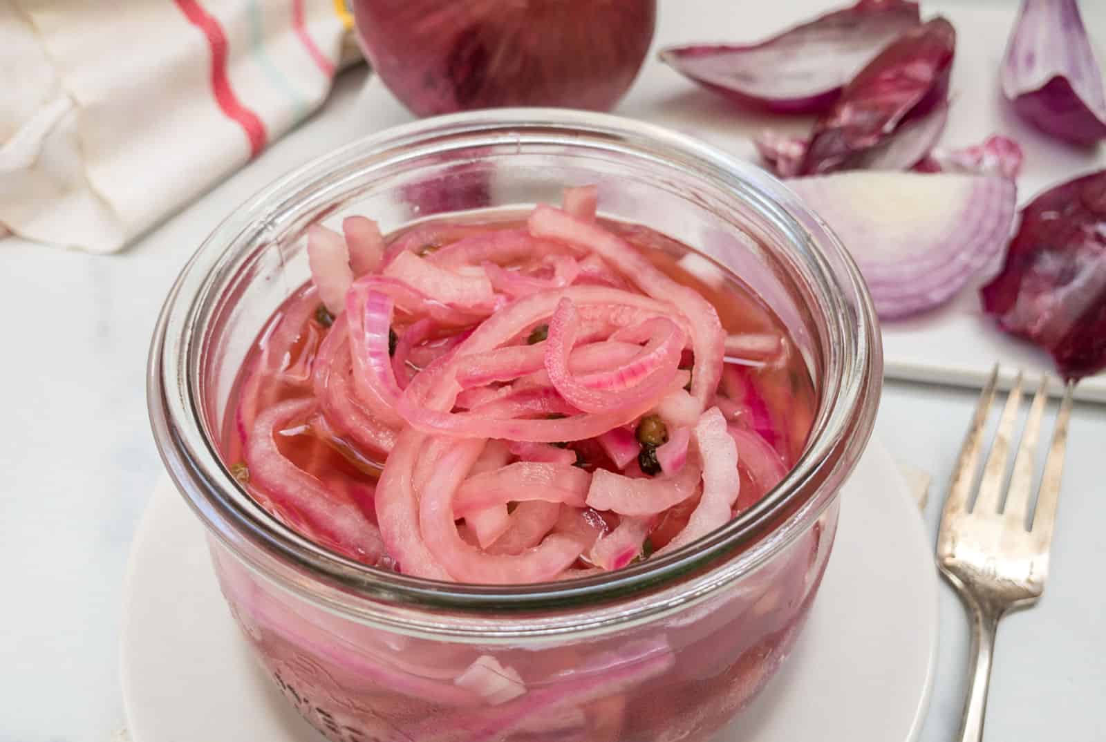 A jar of pickled red onions on a kitchen counter, with raw red onion slices and a fork nearby.