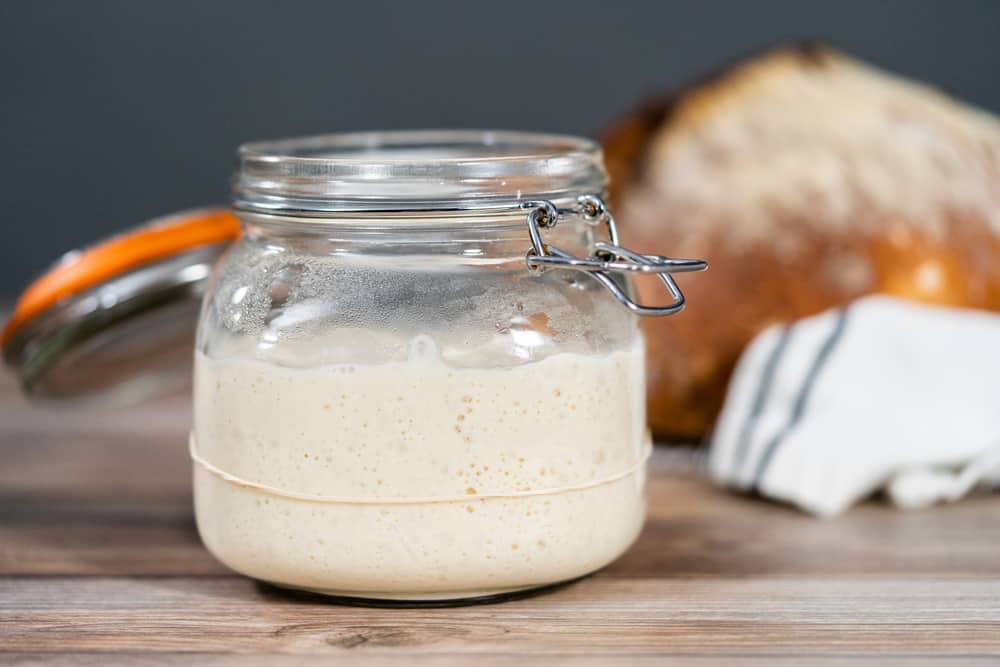 Glass jar containing sourdough starter being fed with a loaf of bread in the background.