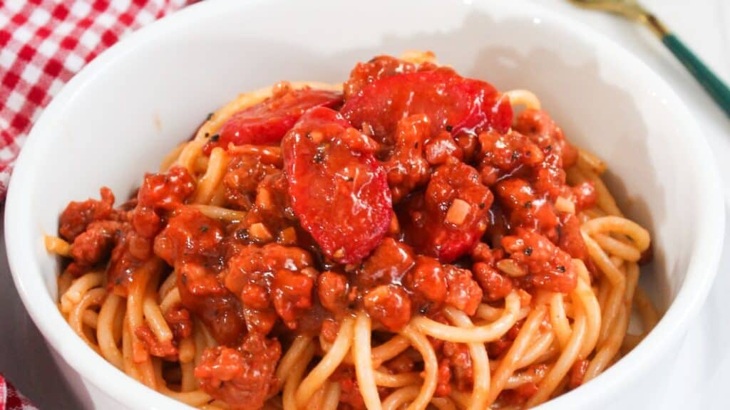 A bowl of spaghetti topped with a rich meat sauce placed on a red and white checkered tablecloth.