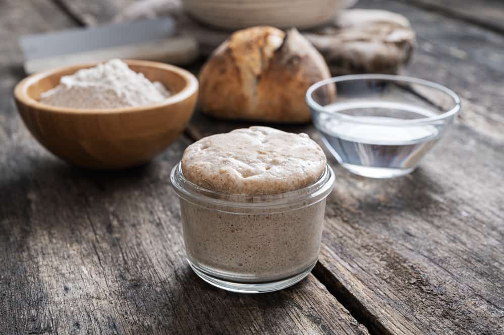 Feeding sourdough starter in a jar with flour, water, and bread on a rustic wooden table.