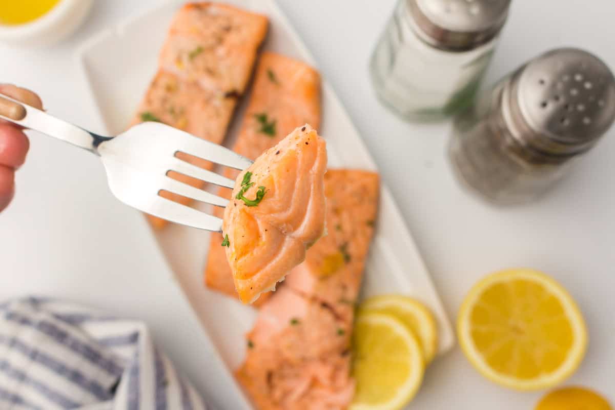 A fork holding a piece of cooked salmon, with more salmon fillets, lemon slices, and seasoning shakers in the background.