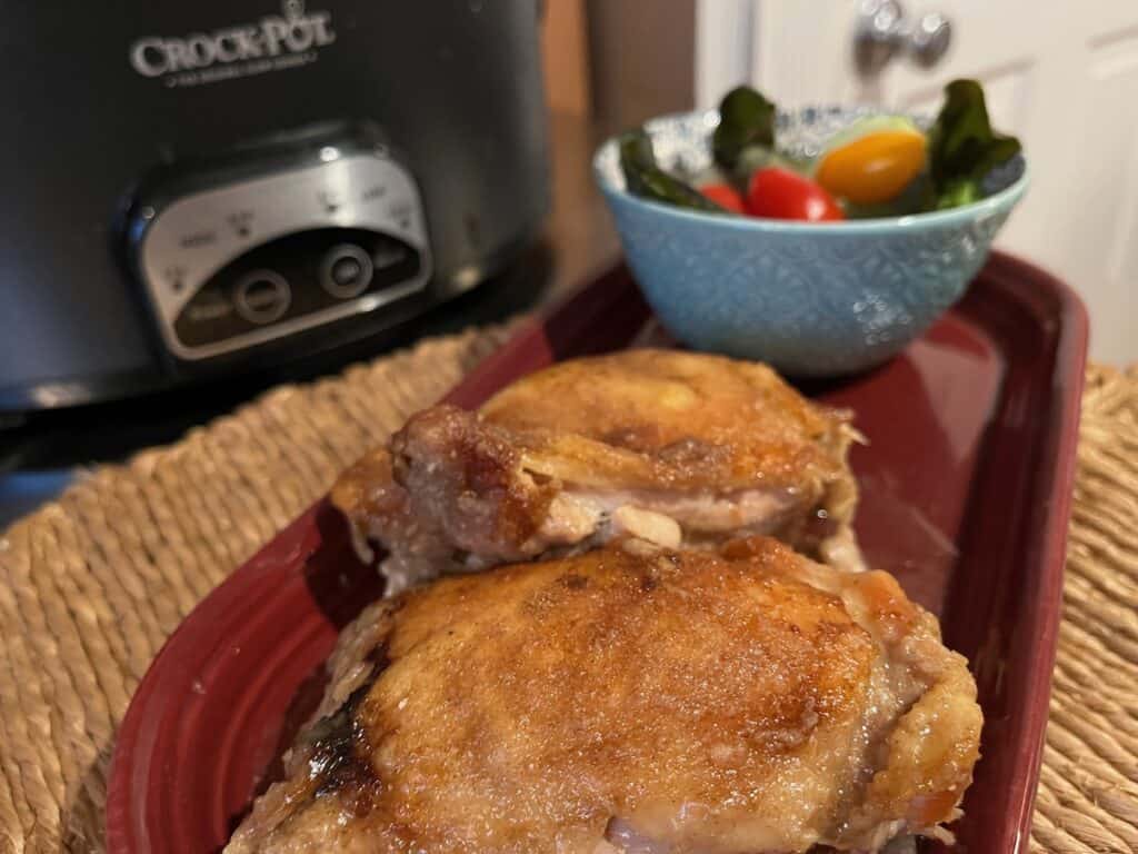 Two cooked chicken thighs on a red dish, a bowl of cherry tomatoes behind, and a slow cooker in the background.