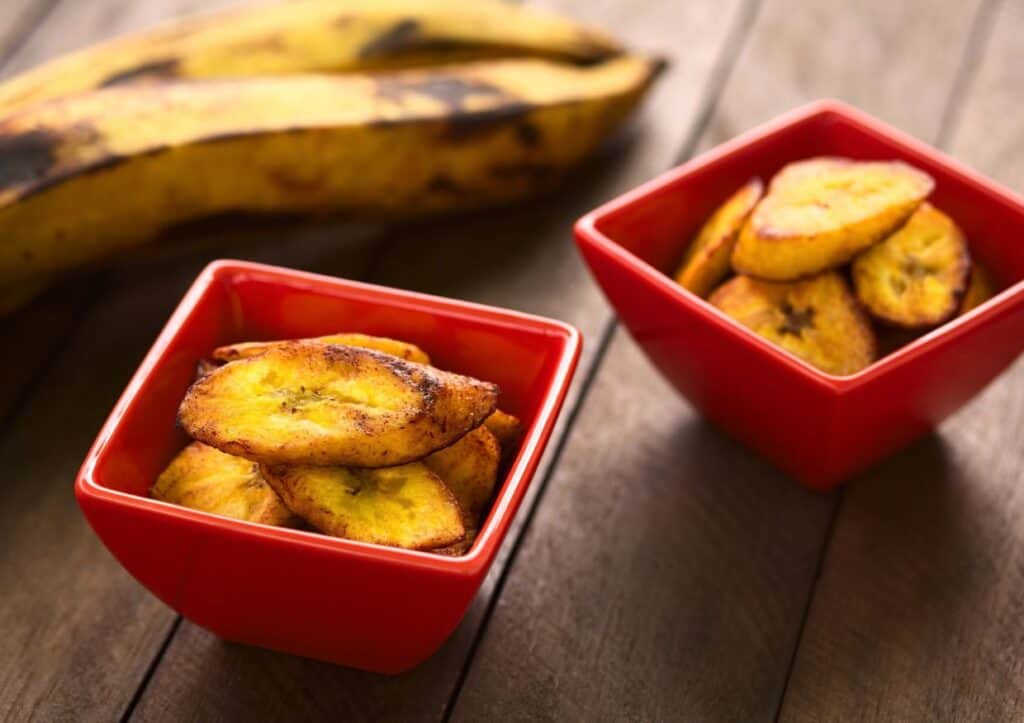 Fried plantain slices in red bowls with a whole plantain in the background on a wooden surface.