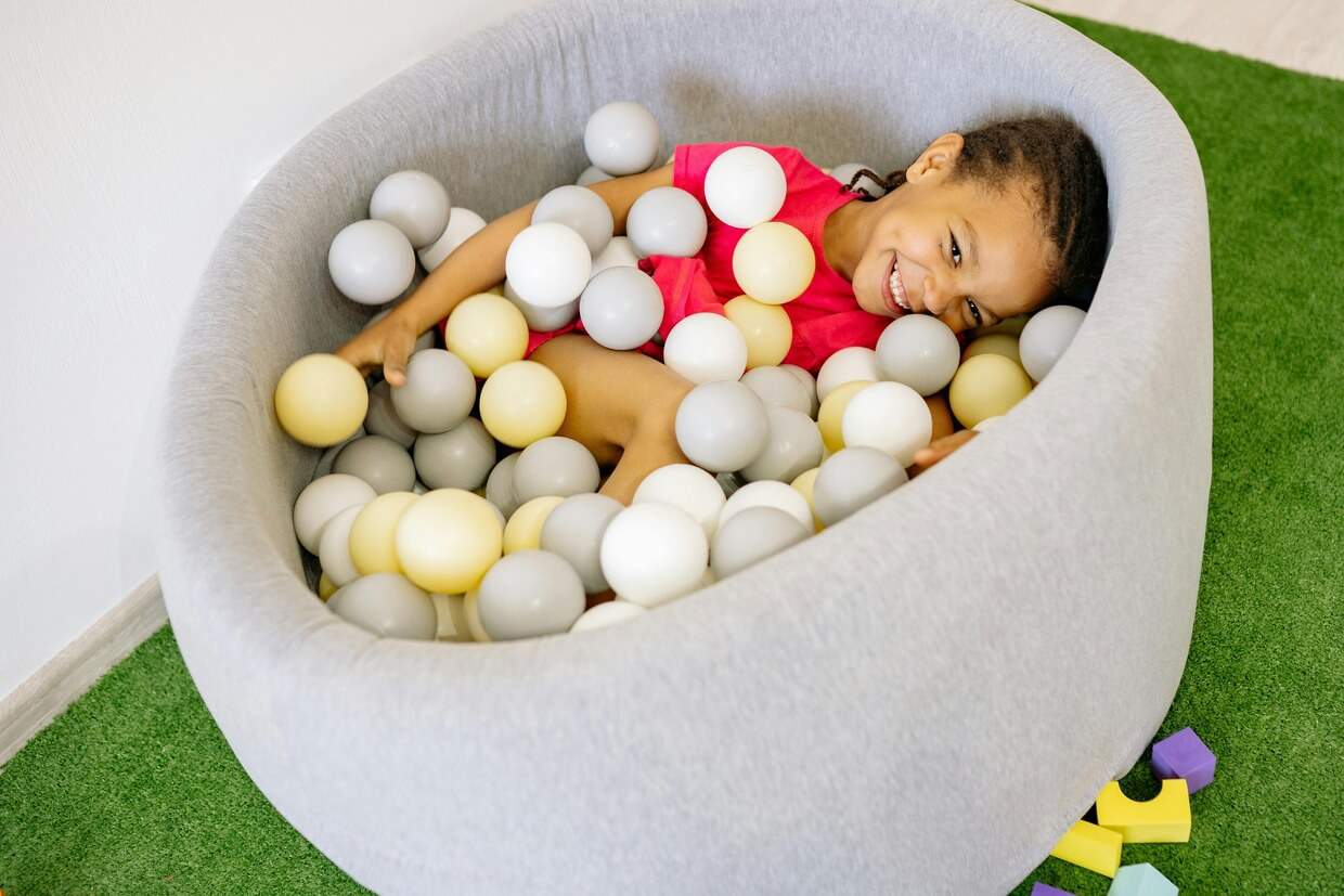 Young child enjoying activities for preschoolers, smiling while lying in a grey ball pit filled with white and beige balls.