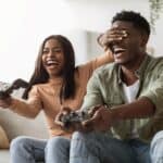 A joyful couple enjoying video games on their date night, with the woman covering the man's eyes playfully on the couch.