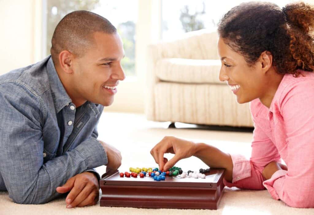 A man and a woman smiling at each other while playing a board game on the floor in a bright living room during their date night.