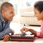 A man and a woman smiling at each other while playing a board game on the floor in a bright living room during their date night.